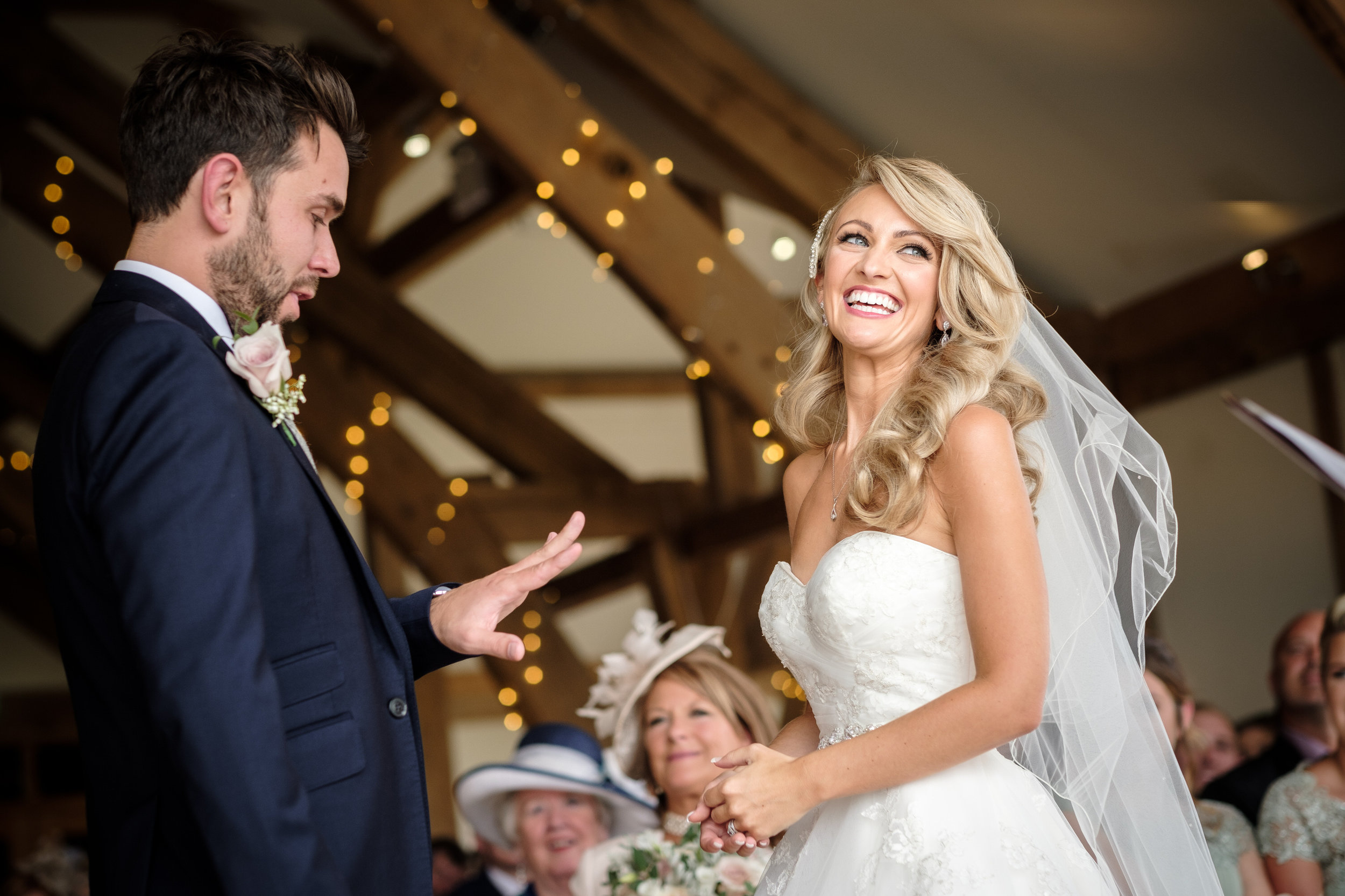 A laughing bride in a strapless gown and veil faces her groom during a barn ceremony lit by fairy lights, guests seated behind them